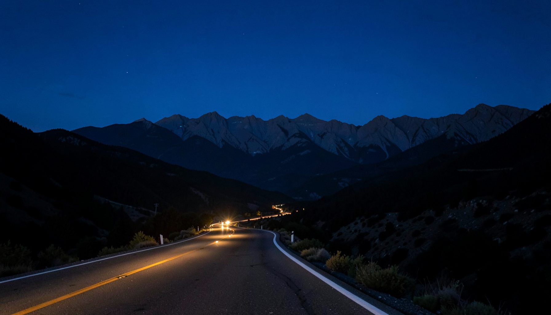 Paisagem noturna com montanhas ao longe sob um céu azul-petróleo profundo, estrada sinuosa iluminada por faróis distantes