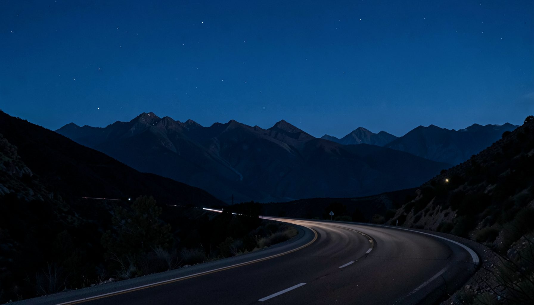 Paisagem noturna com montanhas distantes sob céu azul-petróleo, estrada sinuosa em primeiro plano